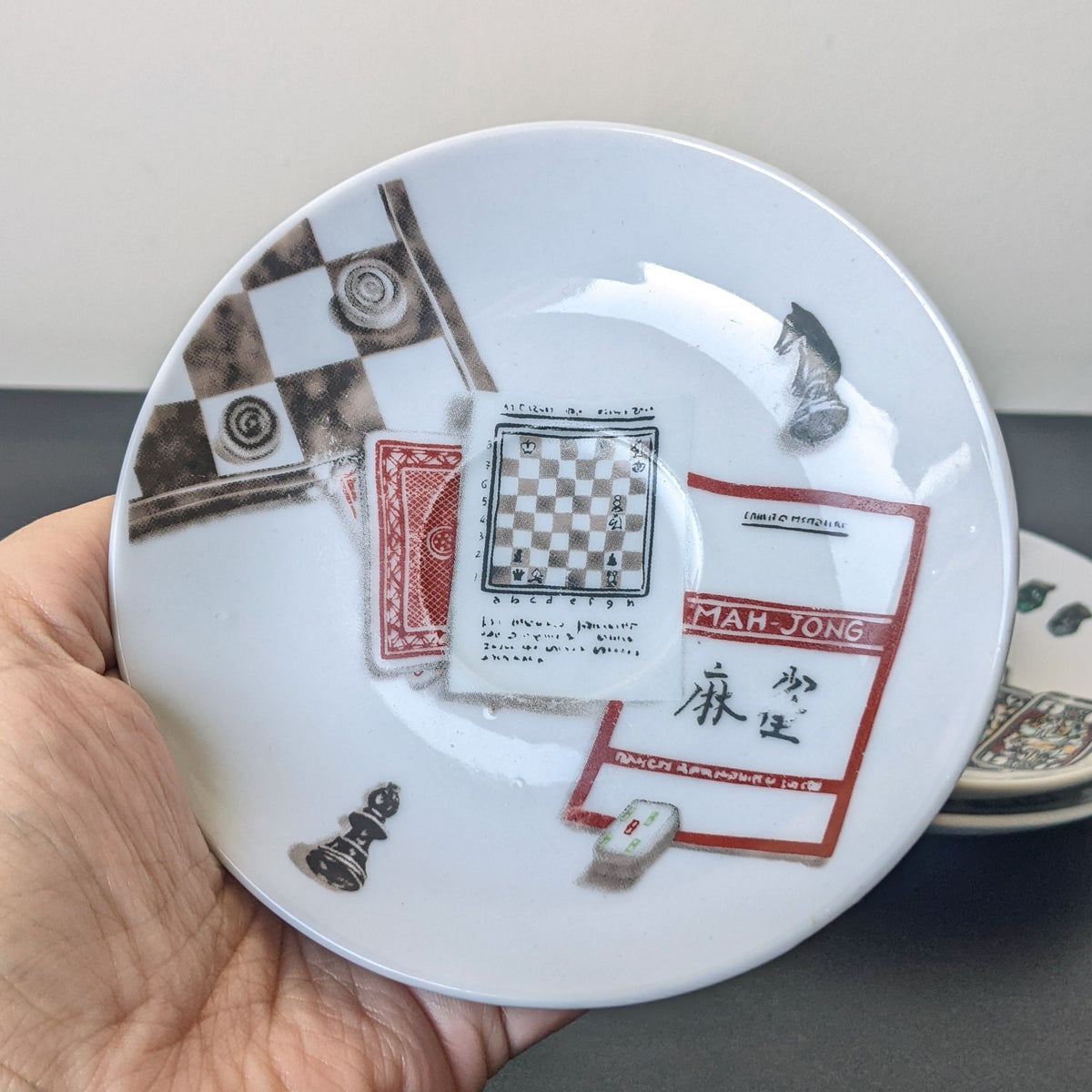 A hand holds a white ceramic plate decorated with chess-themed illustrations, including a chessboard, chess pieces, a playing card, and a red label with Chinese characters. Another matching plate is partially visible in the background on a dark surface.