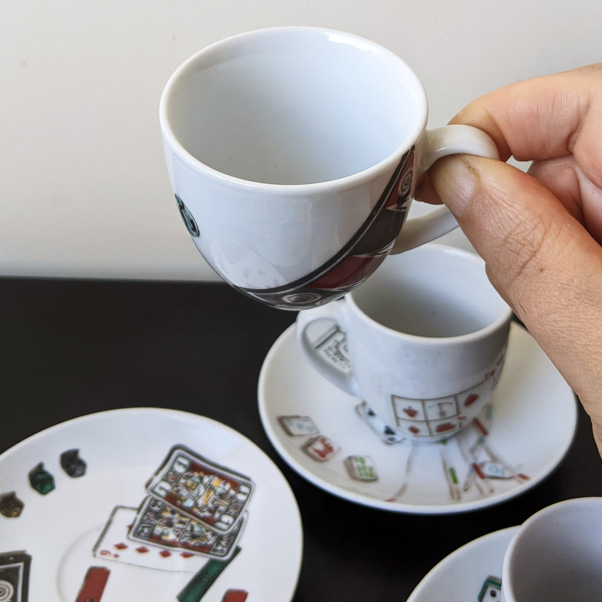 A hand holds a white ceramic espresso cup with a saucer featuring playing card illustrations. Another matching cup and saucer are visible in the background on a dark tabletop against a light wall.